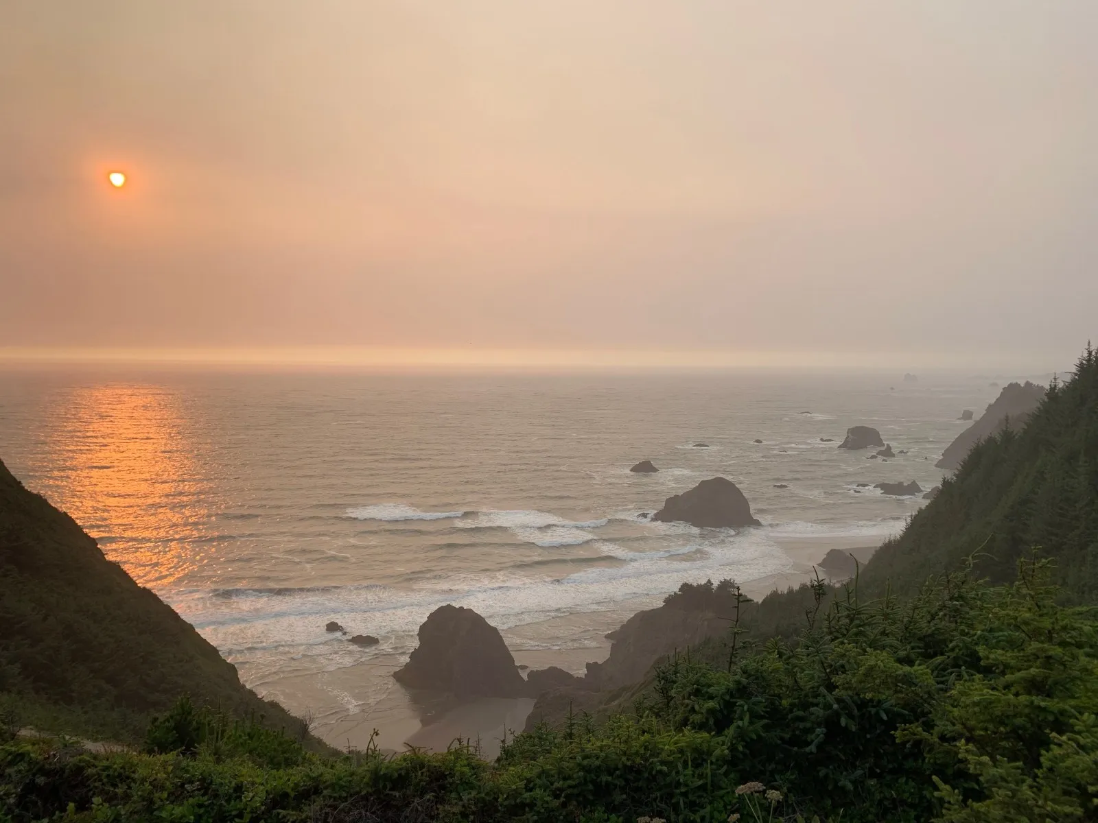 Hazy orange sunset over the Pacific, sea stacks below green cliffs