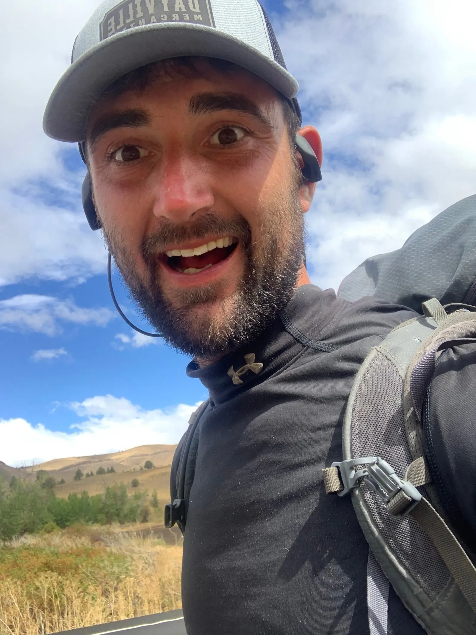Chris grinning in a selfie on the road, cap on, hills behind him